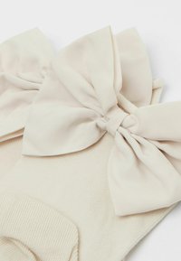 Calcetines beige con textura acanalada, que presentan grandes lazos de tela a cada lado, fabricados con un material suave. Los lazos están atados en el centro.