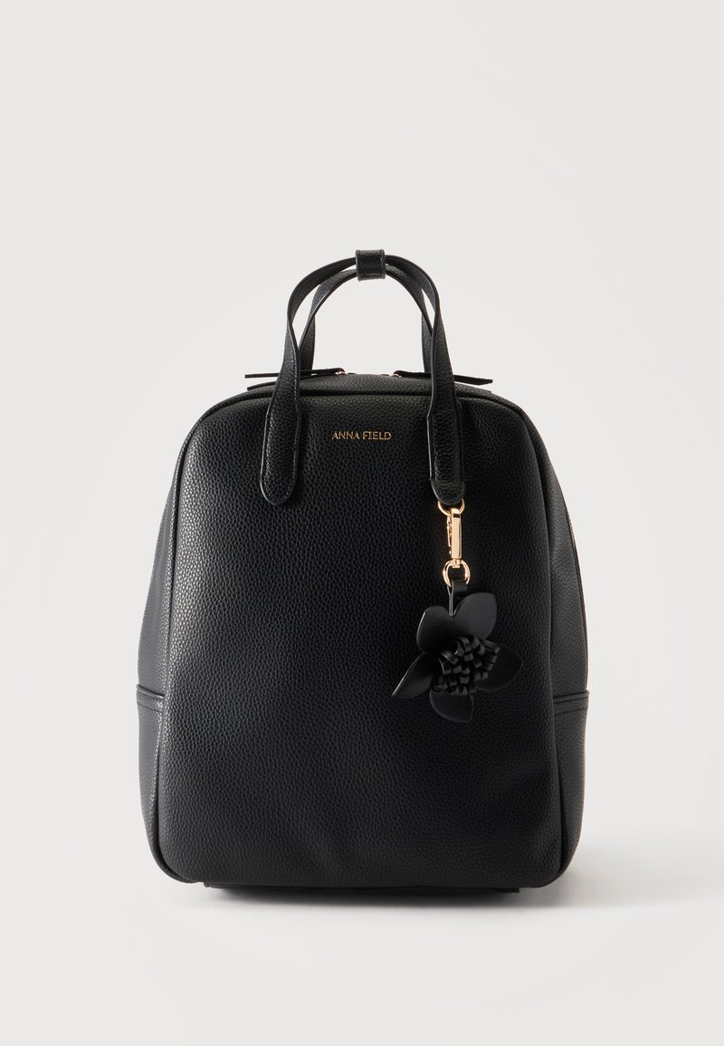 Sac à main en cuir texturé noir avec deux poignées, logo de marque doré et porte-clés fleur noire suspendu à une poignée sur un fond uni.