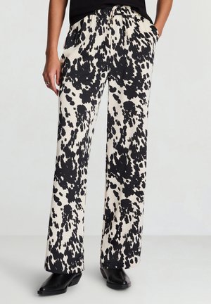 Frau trägt weite, locker sitzende Hosen mit schwarzen und weißen abstrakten Flecken, schwarze Stiefel und ein schwarzes Oberteil, eine Hand in der Tasche.