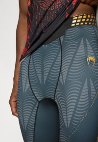 Καλσόν σε σκούρο μπλε με κυματιστό ανοιχτό γκρι σχέδιο, μαύρο ραφές και χρυσή λεπτομέρεια στη μέση. Το κοντομάνικο τοπ φαίνεται μερικώς.