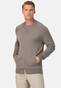 Suéter gris con cremallera, puños y dobladillo acanalados, textura suave, bolsillos laterales y cuello redondo, llevado sobre una camiseta gris de cuello redondo.