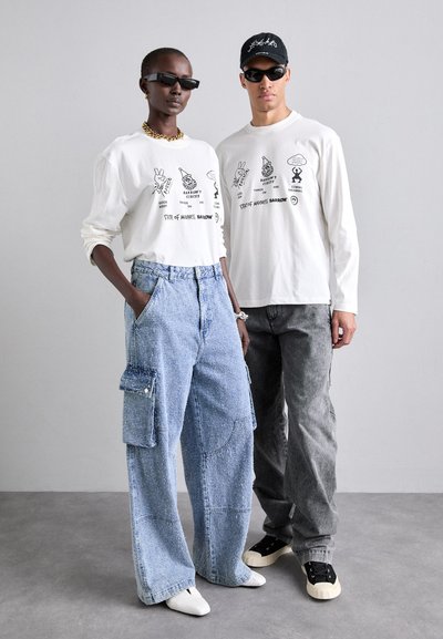 Chemises blanches à manches longues avec des imprimés graphiques, jeans cargo bleu clair avec poches, pantalons gris foncé, lunettes de soleil noires et baskets.