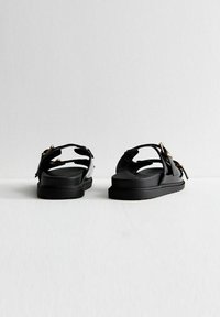 Svarta sandaler med glänsande syntetiska remmar, platta gummisulor och detaljer i guldton. Öppen tå-design med justerbar bakrem.