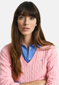 Růžový svetr s pleteným vzorem a výstřihem do V, přes modrou košili s knoflíky. Svetr má římské manžety a malou logovou ozdobu.