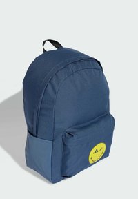 Zaino blu navy con tasca frontale, tasca laterale, manico nero e logo di faccina sorridente che strizza l'occhio gialla sulla tasca, su uno sfondo chiaro.