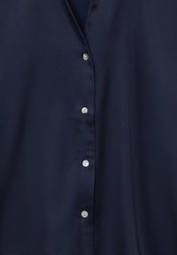 Primo piano di un tessuto blu navy setoso con quattro piccoli bottoni bianchi allineati verticalmente lungo una cucitura.