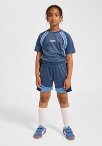 Marinblå träningsoverall med korta ärmar och matchande shorts. Kontrasterande ljusblå detaljer. Ett par blå skosnören, knähöga vita strumpor.