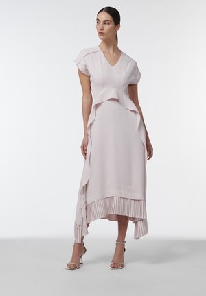 Vrouw staat met een lichtroze, asymmetrische midi-jurk met korte mouwen, geplooide zoom en zilveren hoge sandalen tegen een effen achtergrond.