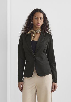 Frau mit lockigem Haar, die einen schwarzen Blazer, einen beige gebundenen Schal um den Hals, ein schwarzes Oberteil und beige Hosen trägt und vor einer weißen Wand steht.