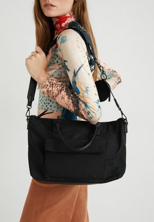 Sac fourre-tout en tissu noir avec deux poignées supérieures, une poche avant et des détails floraux brodés. Sangle réglable attachée. Texture lisse.