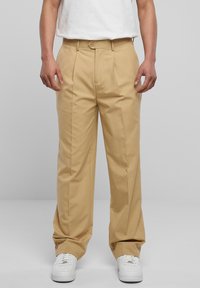 Person som har på seg beige plisserte bukser, hvite sneakers og en hvit skjorte stappet i buksen mot en enkel grå bakgrunn.