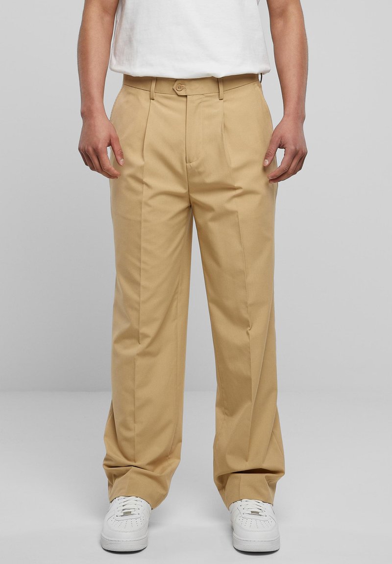 Person som har på seg beige plisserte bukser, hvite sneakers og en hvit skjorte stappet i buksen mot en enkel grå bakgrunn.