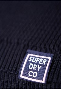 Primo piano di un tessuto nero a coste con un'etichetta testurizzata che riporta "SUPER DRY CO" in carattere bianco bordato da un contorno rosa.