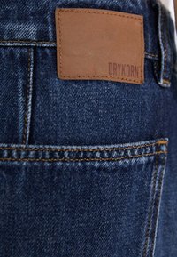 Denim-Jeans mit einer dunkelblauen Waschung, sichtbare Nähte in goldenem Farbton und einem braunen Lederetikett mit eingeprägtem "DRYKORN".