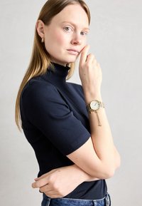Ceas de mână de aur cu cadran alb și brățară din plasă, purtat pe încheietura unui model. Modelul îmbrăcat cu un pulover cu guler înalt, de culoare bleumarin. Fundal luminos.