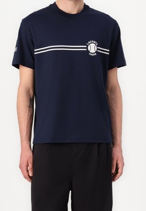Homme portant un t-shirt bleu marine à manches courtes avec des doubles rayures blanches et un logo de balle de tennis "Lacoste Paris" sur la poitrine, assorti à un pantalon noir.