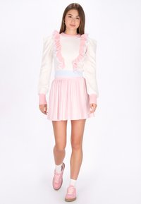 Maglione bianco con dettagli in rouches rosa e blu, abbinato a una gonna plissettata rosa chiaro. La modella indossa scarpe da ginnastica rosa e calzini bianchi alla caviglia.