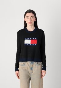 Le modèle porte un sweat-shirt cropped noir affichant le logo "TOMMY JEANS" en blocs de couleur, associé à un jean bleu clair. Fond neutre.