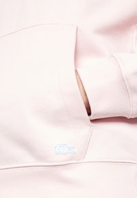 Felpa rosa chiaro con tasca a marsupio e polsini a costine. Presenta un piccolo logo ricamato blu sulla tasca. Materiale in cotone morbido.