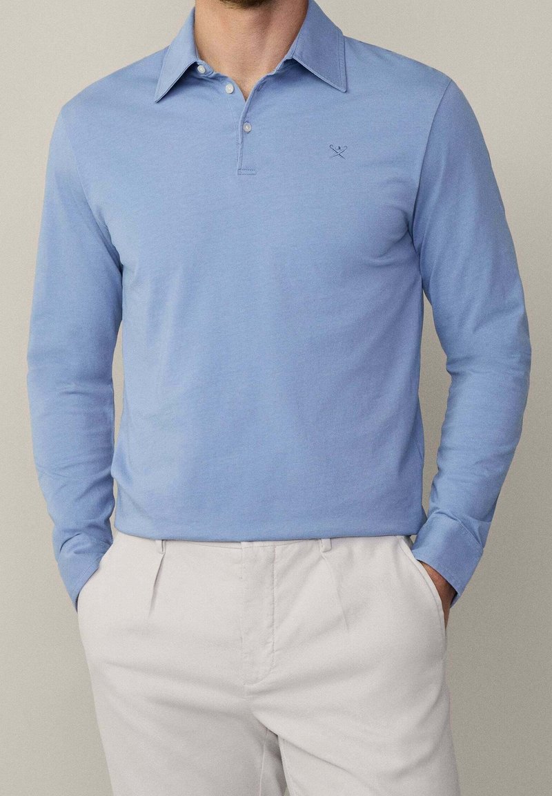 Hellblaues Langarm-Polo-Shirt aus weicher Baumwolle mit einer Drei-Knopf-Leiste und einem kleinen gestickten Detail auf der Brust, kombiniert mit hellen Hosen.