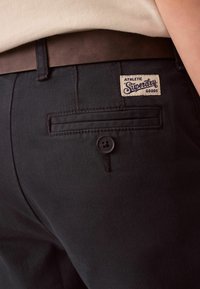 Svarta byxor med slim fit, med en bakficka med knapp och en etikett som läser "Athletic Superdry Goods." Slät textur.