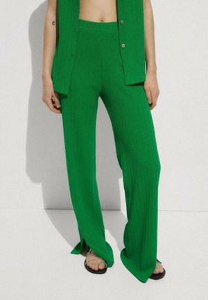 Pantalones anchos de punto acanalado verde con aberturas delanteras, combinados con chaleco sin mangas a juego con botones y sandalias negras de tiras finas.