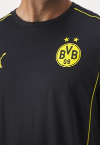 Černé sportovní tričko s žlutým klubovým logem Borussia Dortmund a logem Puma na hrudi, nošené osobou z pohledu od krku po střed trupu.