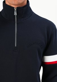 Muž v námořnickém svetru s polovičním zipem, s embosovaným textem "HILFIGER" na hrudi a červenobílým pruhovaným rukávem.