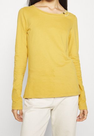 Femme portant un haut manches longues jaune moutarde avec trois petits boutons près de l'épaule gauche et un pantalon beige clair.