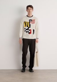 Pull en tricot blanc avec des motifs géométriques colorés et des drapeaux, associé à un pantalon noir et un sac fourre-tout beige.