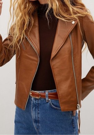 Donna che indossa una giacca marrone in pelle con zip sopra una maglietta nera, jeans blu con cintura in pelle marrone, con lunghi capelli biondi ondulati visibili.