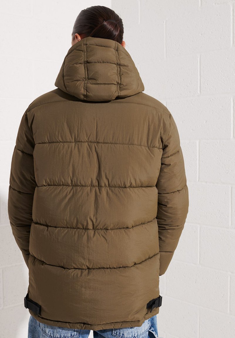 Chaleco de plumas en verde oliva, diseño acolchado, bolsillos frontales, capucha con costuras horizontales, cintas ajustables en el dobladillo y una textura suave.