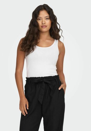 Mujer con cabello largo y rizado, vestida con una blusa blanca sin mangas y pantalones negros de talle alto con un cinturón anudado en la cintura, de pie con una mano en el bolsillo.