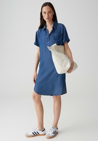 Jeanshemdkleid in Dunkelblau mit kurzen Ärmeln, einer Brusttasche und einem abgerundeten Saum. Kombiniert mit weißen Turnschuhen und einer cremefarbenen Tragetasche.