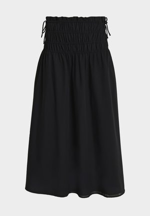 Femme aux longs cheveux foncés portant un haut noir sans manches avec peplum et une jupe midi à larges jambes, tenant une pochette noire et portant des talons noirs.