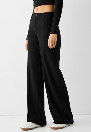 Pantalons noirs à jambes larges avec une texture lisse, dotés d'une fermeture à bouton à l'avant et de plis frontaux subtils, associés à des chaussures blanches.
