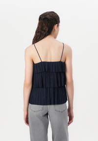 Débardeur plissé bleu marine avec trois volants superposés, fines bretelles réglables et ourlet droit. Porté avec un jean large gris clair.