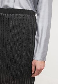 Svart plisserad kjol med texturerade remsor och elastisk midja, kombinerad med en ljusgrå satin långärmad skjorta som visar en kontrast av slät tygstruktur.