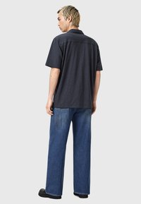 Tummansininen lyhythihainen paita kauluksella, yhdistettynä väljiin sinisiin farkkuihin ja mustiin kenkiin, nähtynä takaa.