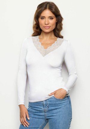 Camicia bianca a maniche lunghe con scollatura a V rifinita in pizzo, morbida al tatto e dalla vestibilità aderente, abbinata a jeans in denim blu.