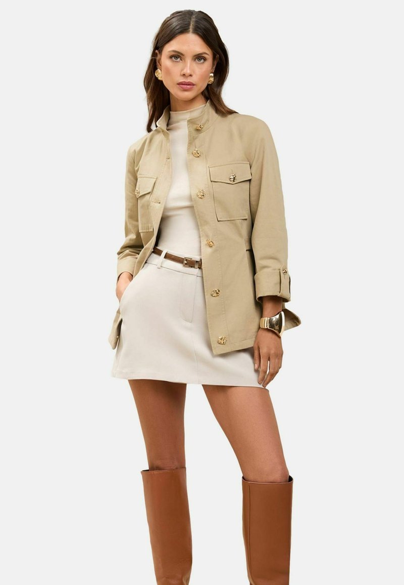 Beige jacka med guldknappar, två bröstfickor, tillsammans med en ljusfärgad minikjol och bruna knähöga stövlar. Slät textur, strukturerad passform.