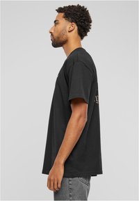 Juodas medvilnės marškinėliai su trumpomis rankovėmis, laisvo kirpimo, su grafiniu spaudiniu priekyje ir gale, demonstruojami ant modelio šone.