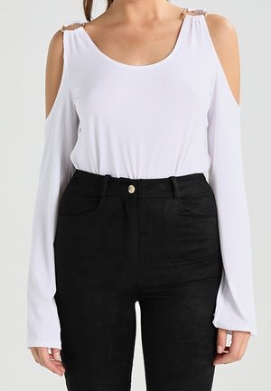 Vrouw draagt een witte cold-shoulder top met lange mouwen en ringdetails en een zwarte broek met hoge taille tegen een effen achtergrond.