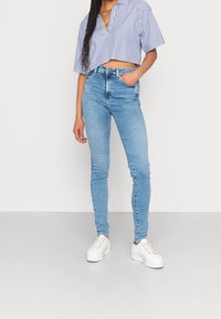 Joven con jeans ajustados de color azul claro, zapatillas de plataforma blancas y una camiseta corta a rayas azul y blanca, de pie con una mano en el bolsillo.