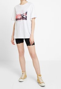 Vit bomulls T-shirt med tryckt grafik, ihop med svarta shorts med spetsdetaljer och bruna hög-top sneakers.