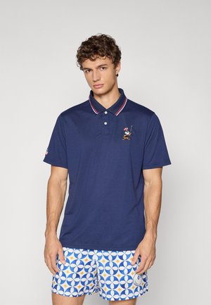 Marineblå polo-shirt med en rød og hvid stripet krave, brodert golferlogo på brystet, i kombination med mønstrede blå og hvide shorts.