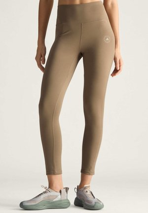 Brune højhalsede leggings med en glat tekstur, der har et lille logo på hoften, sammen med grå og grønne træningssko.