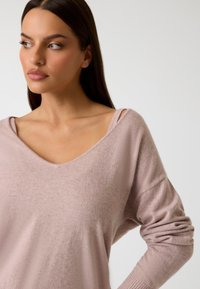 Suéter suave de un ligero tono rosa con un escote en forma de amplia v y estilo de hombros al descubierto. Cuenta con mangas largas con puños acanalados y una textura suave.