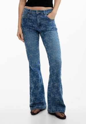 Kobieta ubrana w niebieskie dzwony z białymi kwiatowymi wzorami, czarne kabaretki, czarne buty z odkrytymi palcami oraz czarny krótki top.