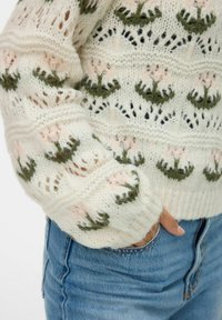 Pull en tricot crème avec un motif floral vert et rose. Présente des mailles ouvertes et des poignets côtelés, associé à un jean en denim bleu.
